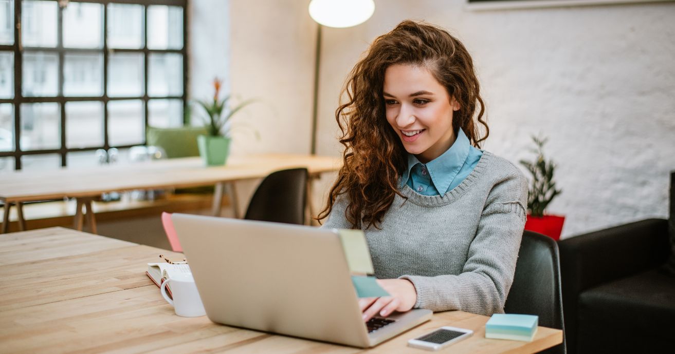 Woman emailing in home office using laptop with phone on desk